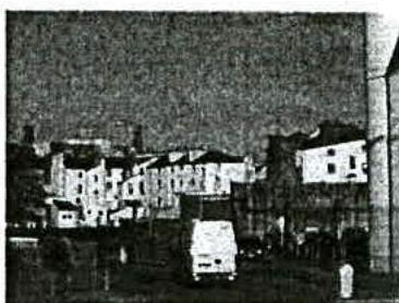 Grainy black and white photograph depicting a street scene with multi-story buildings and a white van in the foreground.