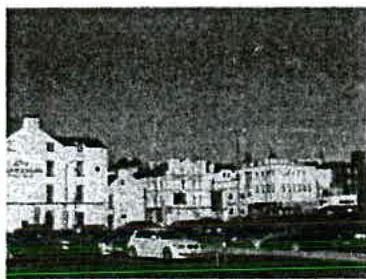 A grainy, low-resolution photograph showing a row of buildings along a street with cars parked in the foreground.