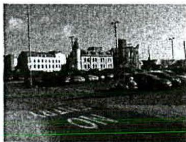 Grainy black and white photograph showing a street scene with parked cars and multi-story buildings in the background.