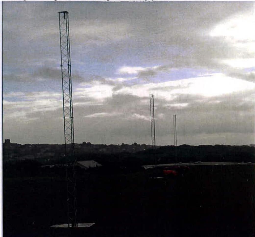 A photograph showing three tall lattice masts silhouetted against a bright, cloudy sky, likely illustrating the proposed telecommunications infrastructure.