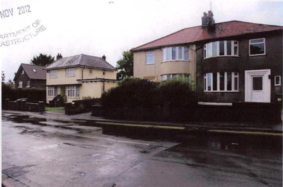A street-level photograph showing existing residential properties, including a cream-colored detached house and a larger two-story house with a red roof, likely the site of the proposed demolition.