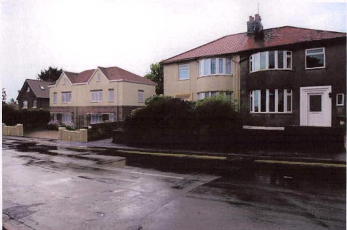 A street-level photograph showing a row of existing residential properties, including a detached cream-colored house and a larger two-story home with dark cladding, likely representing the site context.