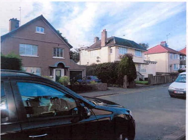A street-level photograph showing a row of existing residential houses, including a brown brick house and a cream-colored house, with a black car in the foreground.