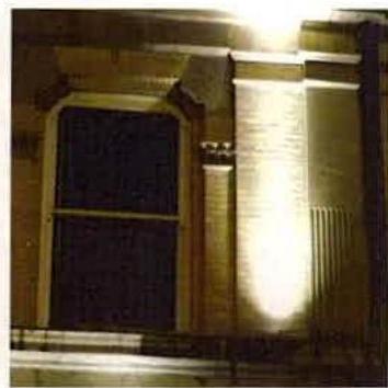 A close-up photograph of a building facade at night, showing a sash window and a wall illuminated by a bright external light fixture.