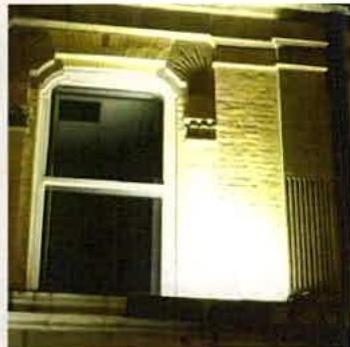 A close-up night photograph of a brick building facade illuminated by an external light source. The image highlights a window and part of a boundary fence.
