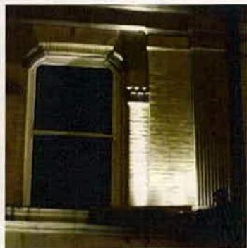 A night-time photograph showing external uplighting on a stone building facade, highlighting an arched window and a vertical column.