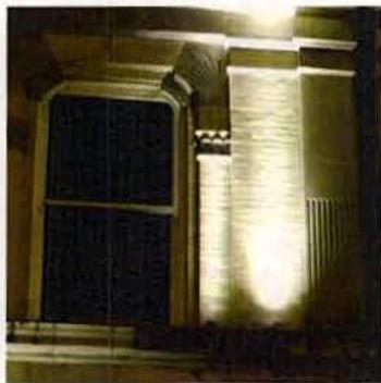 A night-time photograph showing the exterior facade of a building, specifically highlighting a window and a column illuminated by artificial lighting.