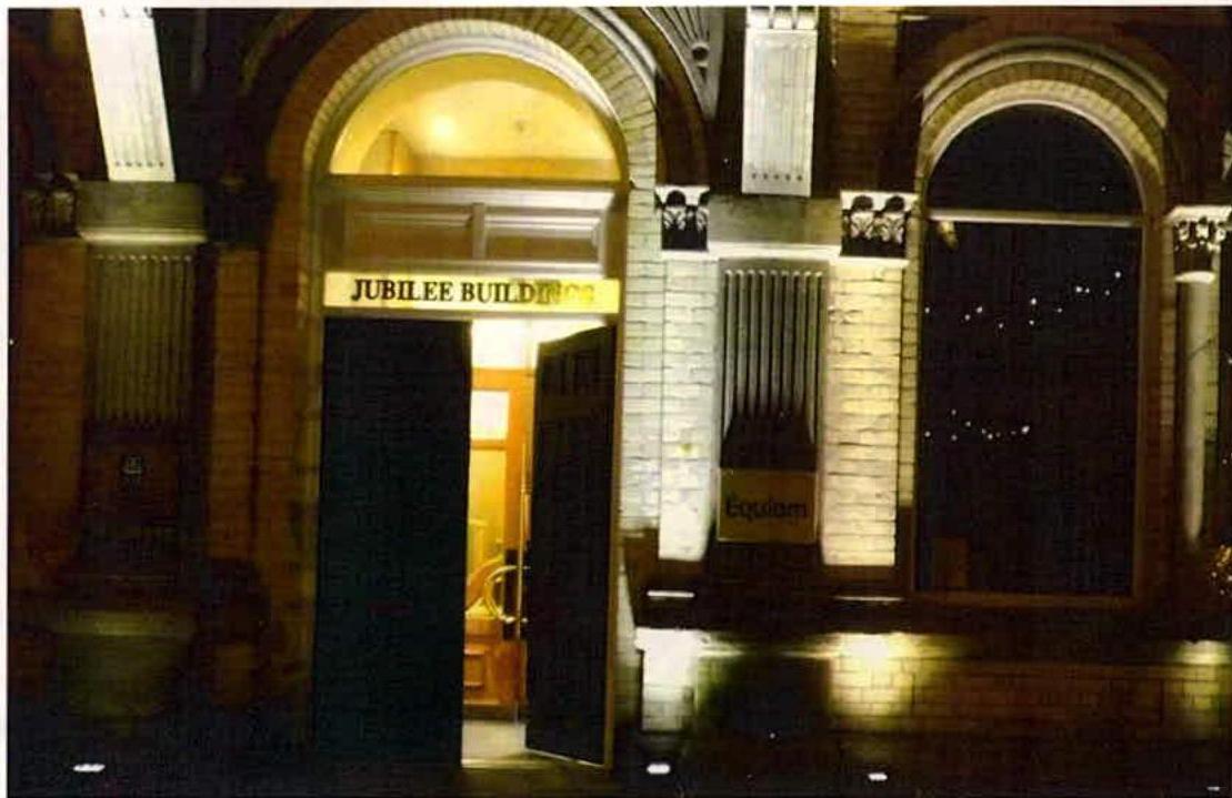 A night-time photograph of the Jubilee Buildings facade, showing the illuminated entrance archway and brickwork.