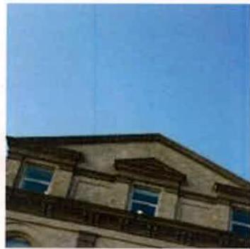 A low-angle photograph showing the upper facade of a stone building with windows and a balcony railing against a clear blue sky.