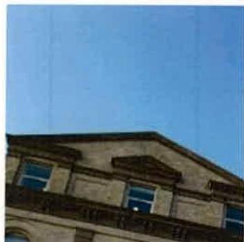 A low-angle photograph showing the upper facade and roofline of a stone building with classical architectural details against a clear blue sky.