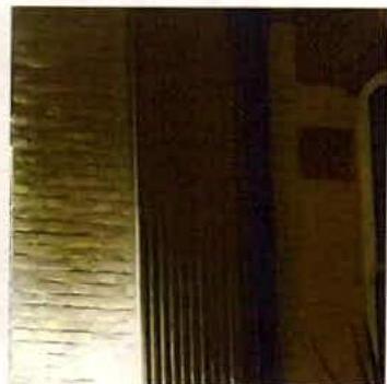 A grainy, low-light photograph showing a close-up of an illuminated exterior wall surface, likely demonstrating the proposed lighting. The image captures a textured wall on the left and a darker section to the right.