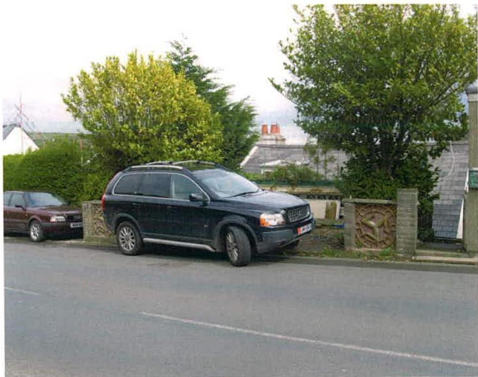 A street-level photograph showing parked cars on a road with residential houses and trees in the background.