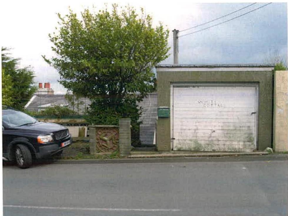 A street-level photograph showing a detached garage with a white roller door and a large tree, with a parked car on the left and a neighboring house roof visible in the background.
