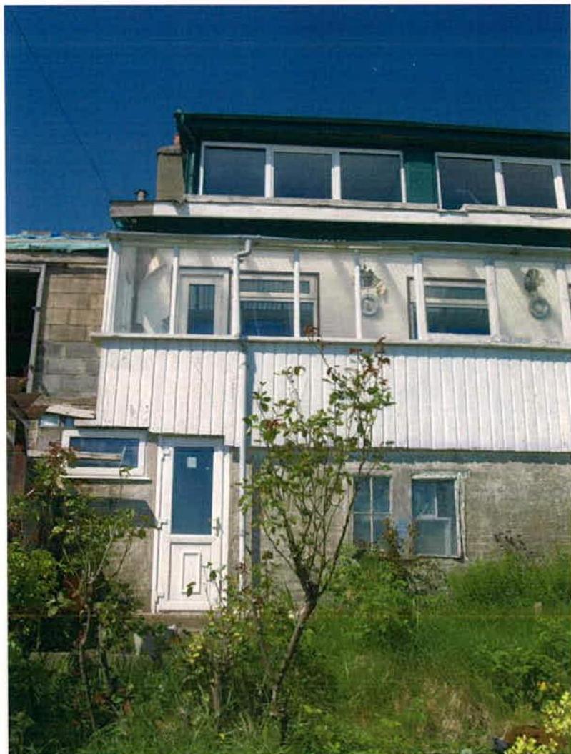 A photograph of an existing detached house featuring white cladding and a flat roof extension, surrounded by overgrown vegetation.