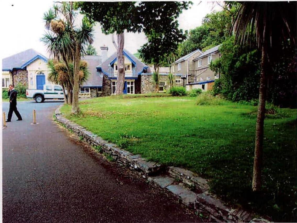 A street-level photograph showing a paved road, a large grassy lawn, and stone buildings with blue trim in the background.