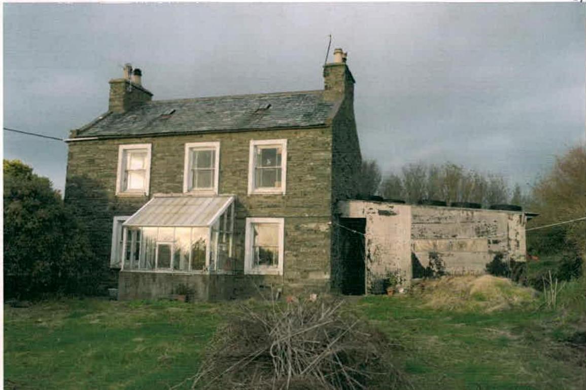 A photograph of a two-story stone dwelling featuring a conservatory and an adjacent dilapidated outbuilding in a rural setting.
