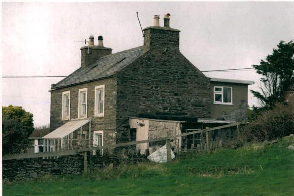 A photograph of a two-story stone detached house featuring existing single-story extensions and a conservatory, situated in a rural setting.