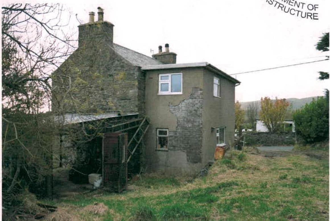 A photograph of a rural two-storey dwelling featuring a mix of stone and rendered walls with an attached lean-to structure.