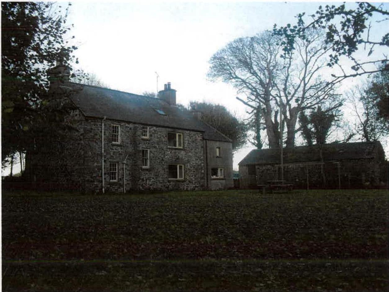 A photograph showing a two-story stone dwelling with a slate roof and a smaller stone outbuilding to the right, set in a rural environment with trees and grass.