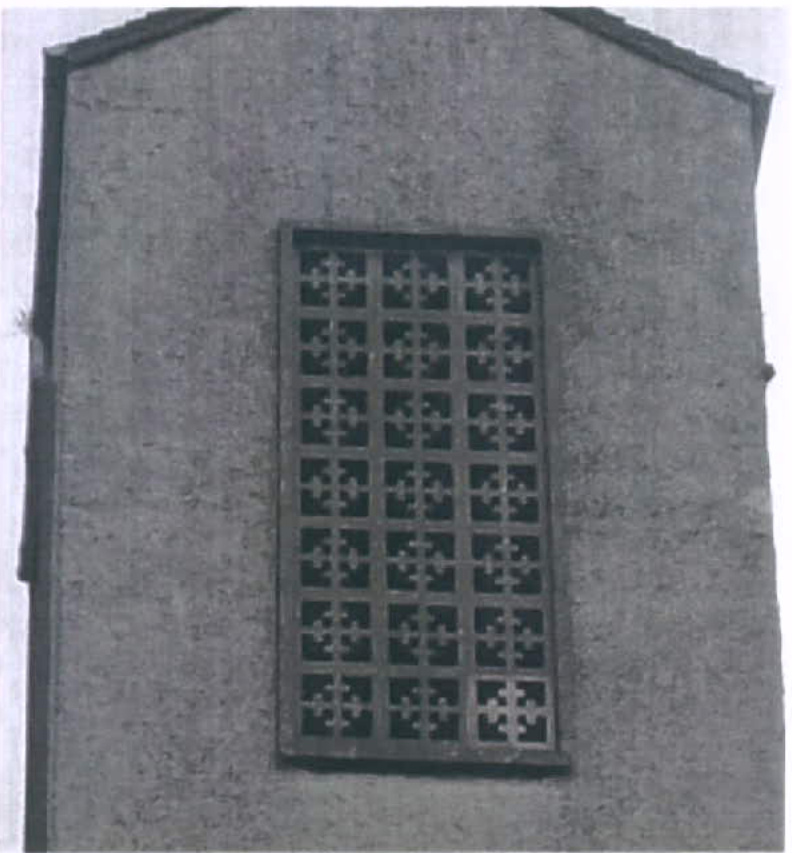A black and white photograph showing a close-up of a textured grey wall with a large rectangular metal grille featuring a repeating cross pattern.
