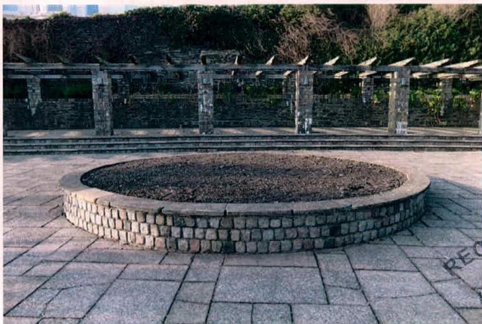 A photograph of a paved public space featuring a circular stone-edged planter bed in the foreground and a stone retaining wall with a wooden pergola in the background.