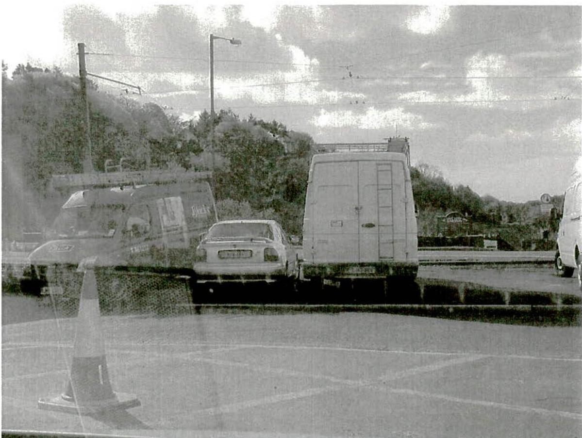 A grainy black and white photograph showing a parking lot with a car and a large van parked near a tree-lined slope.