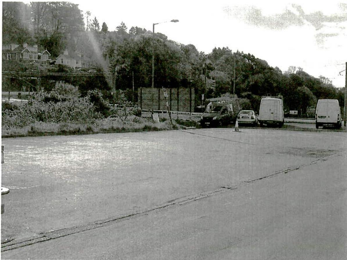 A black and white photograph showing a large paved area with several vehicles parked near a fence, with houses visible on the hillside in the background.