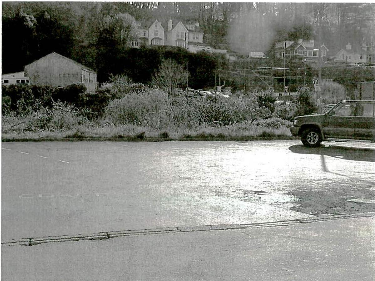 A black and white photograph showing a paved surface, likely a parking area, with a vehicle parked on the right and houses on a hillside in the background.