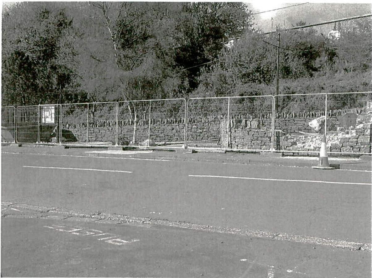 Black and white photograph showing a roadside area with temporary metal fencing, a stone wall, and vegetation in the background.