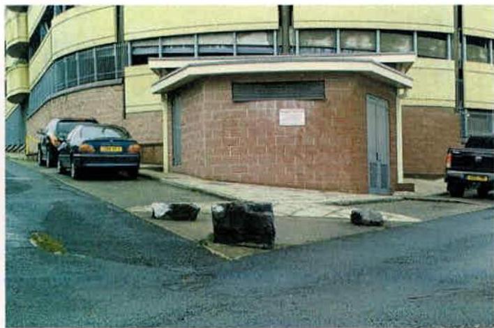 A street-level photograph showing a large curved commercial building with yellow cladding and a smaller brick structure in the foreground with parked cars nearby.