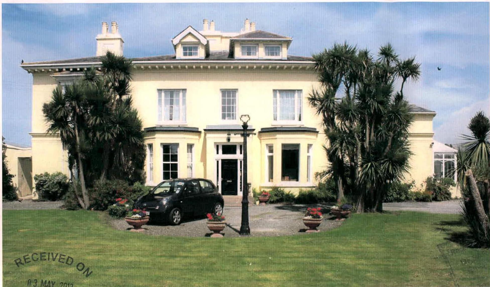 A photograph showing the front elevation of a large, detached, cream-colored house with a gravel driveway, parked car, and palm trees.