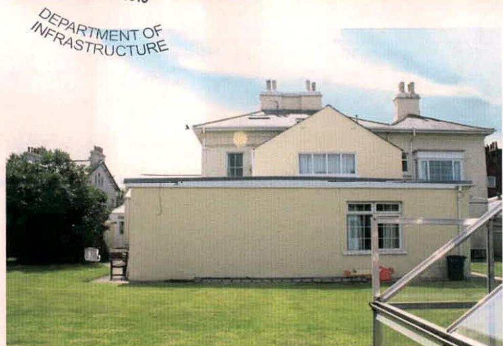 A photograph showing the rear elevation of a cream-colored detached house featuring a single-story extension and a greenhouse in the garden.