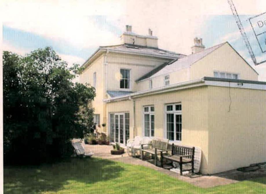 A photograph showing the rear or side elevation of a large, cream-colored detached house with a single-story extension, featuring a patio area with benches and a lawn.