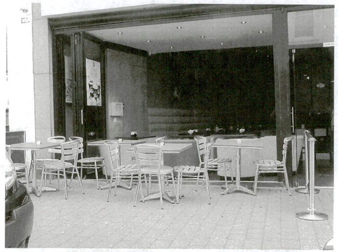 Black and white photograph showing the exterior of a commercial property with tables and chairs arranged on the pavement outside.