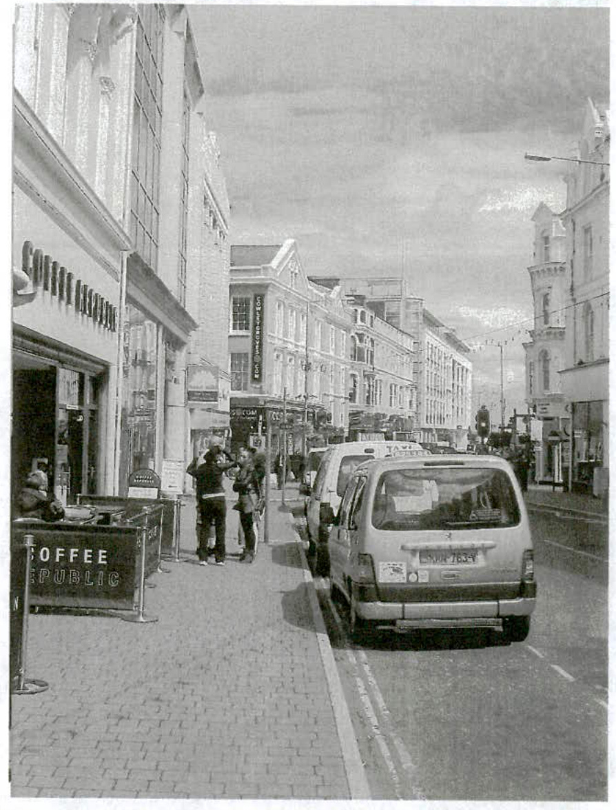 A black and white photograph showing a street scene with a 'Coffee Republic' storefront and metal pavement barriers for outdoor seating.