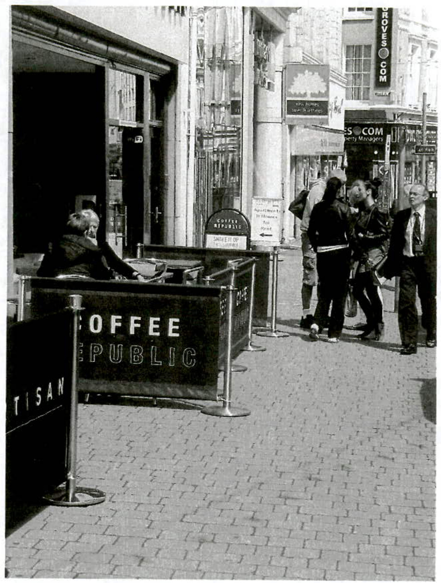 Black and white photograph showing a street scene with pavement barriers branded 'Coffee Republic' enclosing an outdoor seating area. Pedestrians are walking on the sidewalk while people sit at tables behind the barri...