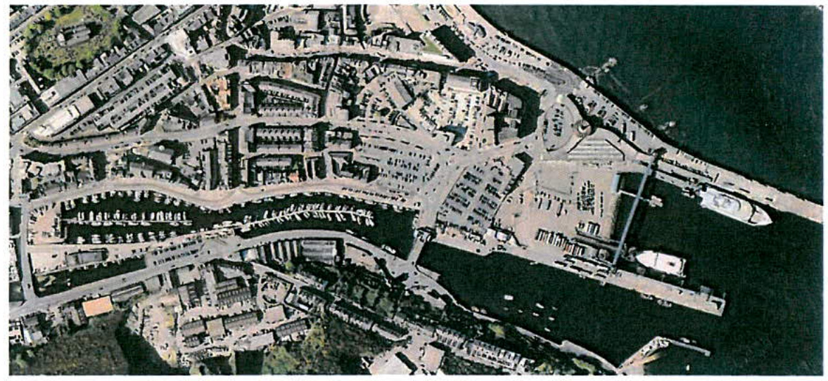 High-angle aerial view of a coastal harbor area featuring boats, parking lots, and surrounding town buildings.