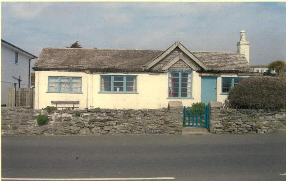 A front view photograph of a single-storey detached bungalow with cream render, slate roof, and a stone boundary wall.