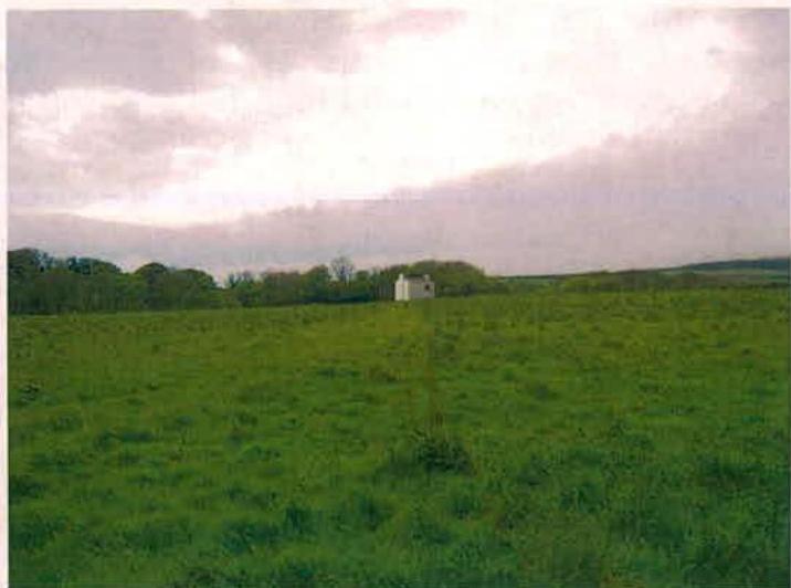 A photograph showing a rural landscape with a large green field in the foreground and a small white cottage visible in the distance near a tree line.