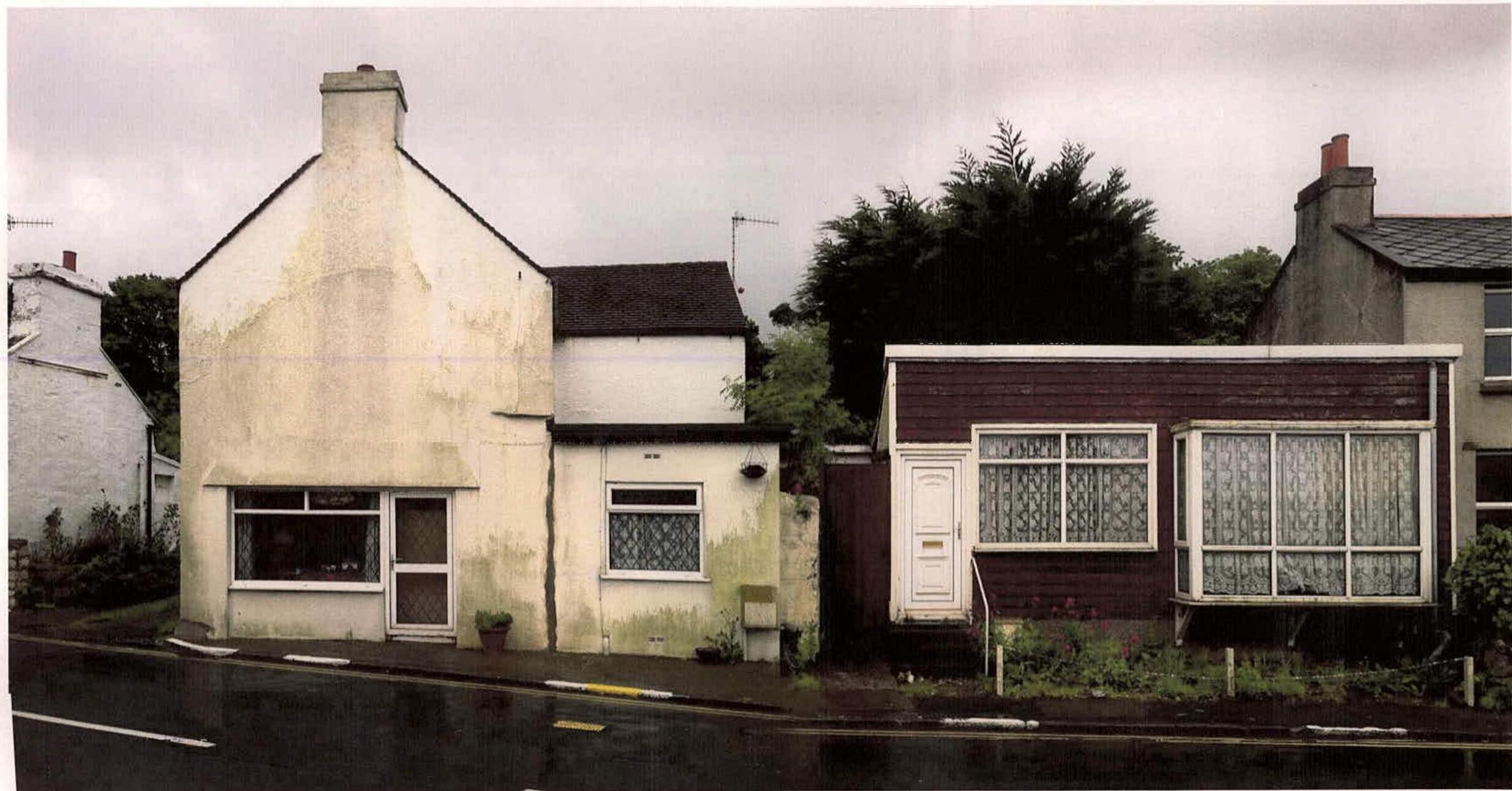 A street-level photograph showing a white rendered house with a pitched roof adjacent to a single-story dark-clad structure with a flat roof.
