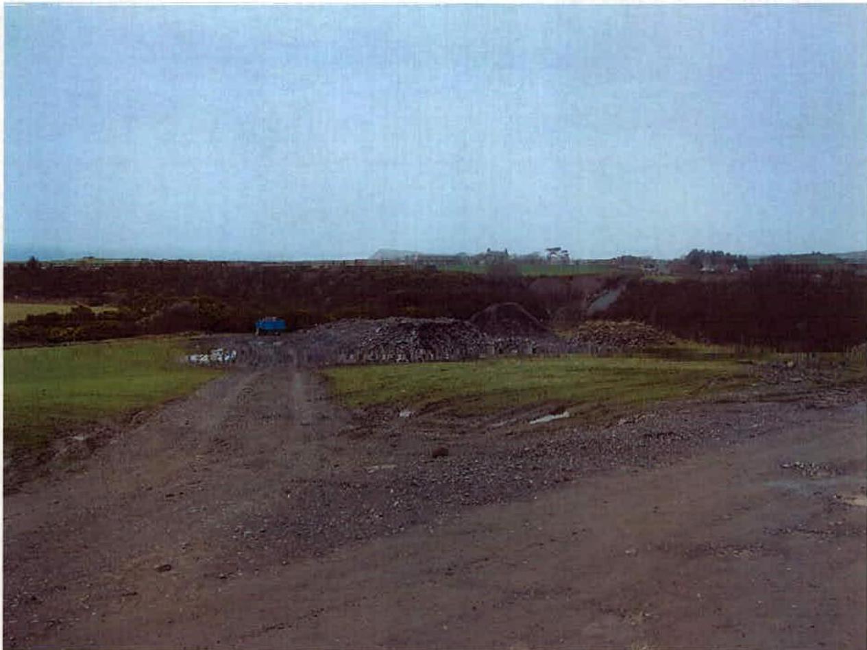 A photograph of a rural site featuring a dirt track leading towards a pile of rubble or stone, with hedgerows and distant buildings in the background.