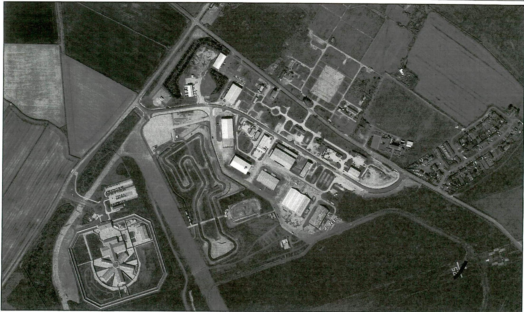 Black and white aerial photograph showing a rural site featuring a winding go-kart track, several large industrial buildings, and surrounding agricultural fields.
