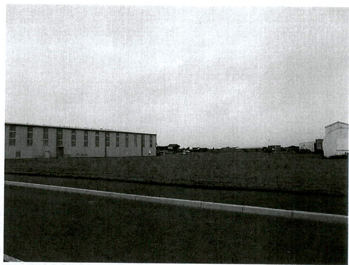 A grainy black and white photograph showing a long single-story building on the left and a grassy field with a paved road in the foreground.