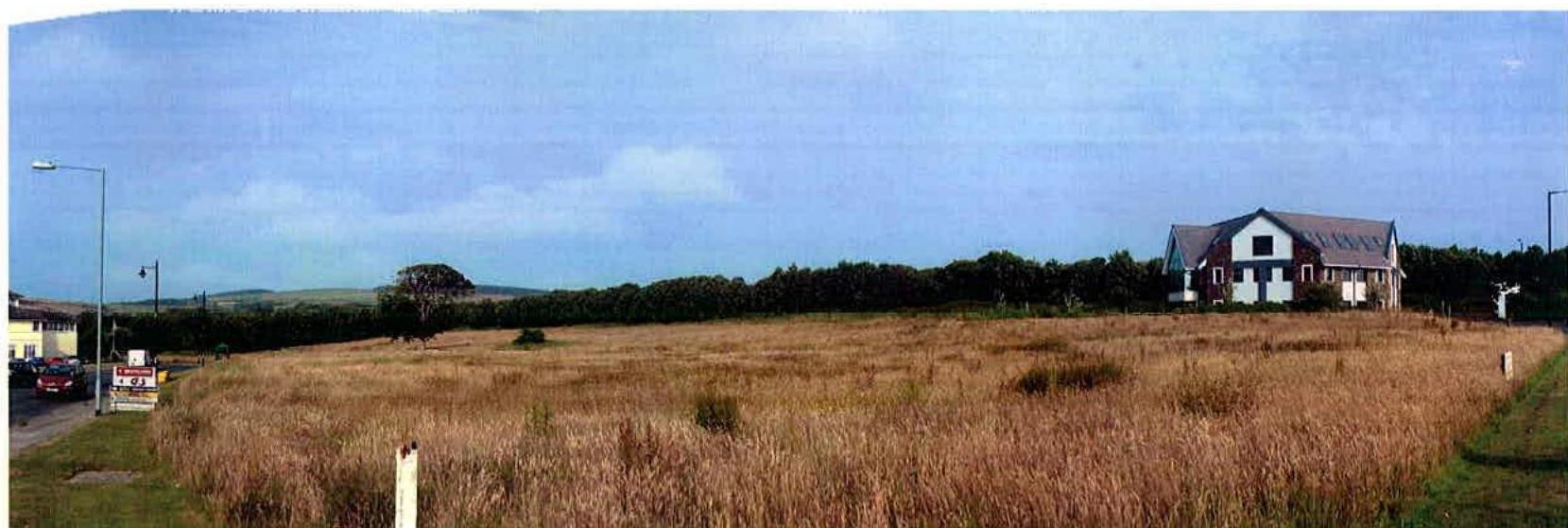 A wide landscape photograph showing a large, grassy field in the foreground with a large white house in the background on the right and a road on the left.