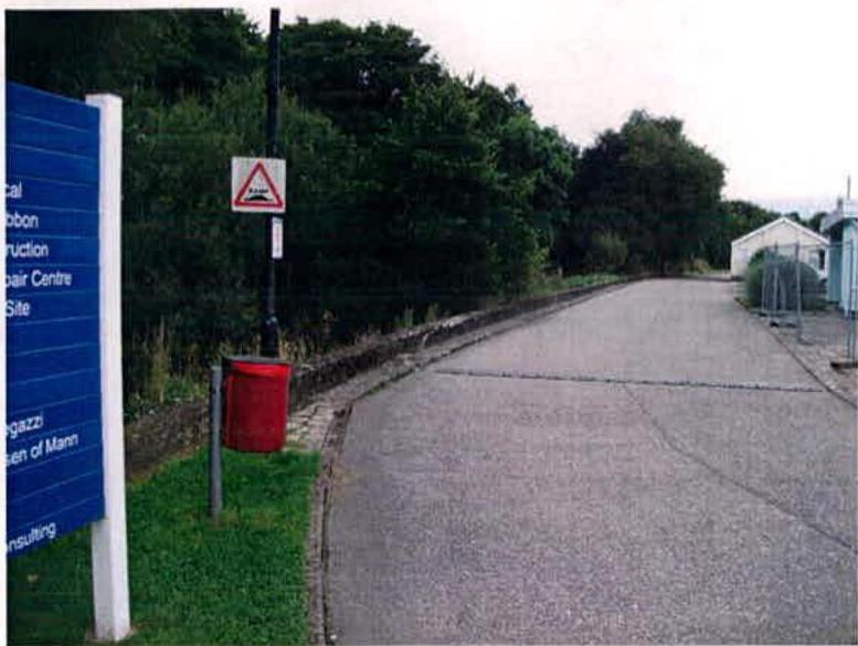 A photograph showing a paved access road with a blue directory sign on the left and white buildings in the distance.
