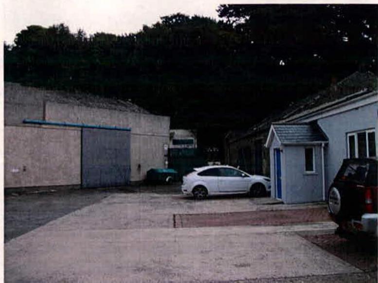 A photograph showing a paved driveway area with parked cars, flanked by a large concrete building with a blue door on the left and a smaller white outbuilding on the right.