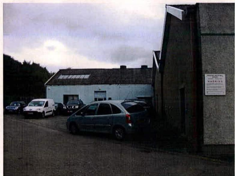 A photograph showing a parking area with several cars in front of a light-colored building with a pitched roof and skylights. A sign is visible on the corner of a building in the foreground.