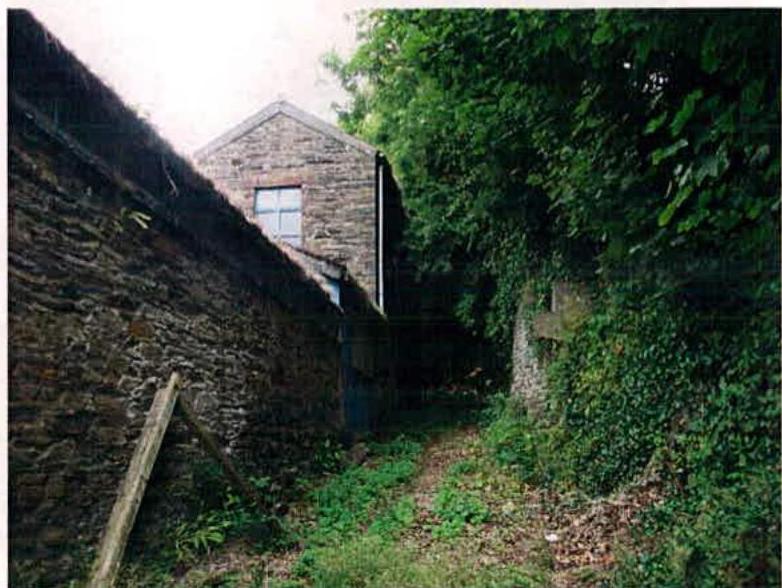 A photograph showing a stone building viewed from a narrow path, flanked by a stone wall on the left and dense vegetation on the right.