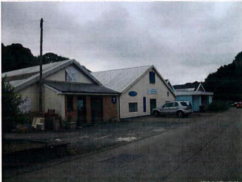 A photograph showing a row of single-story buildings, likely commercial or community use, with a car parked in front. One building on the left appears to be in a state of disrepair or renovation.