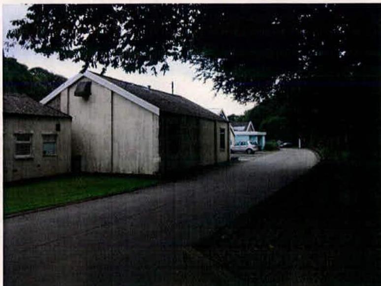 A street-level photograph showing a long, single-story white building with a pitched roof situated next to a road and trees.